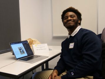 Shawn Patterson-Lee sitting by a laptop with his project, "BMO: Black Magnum Opus."